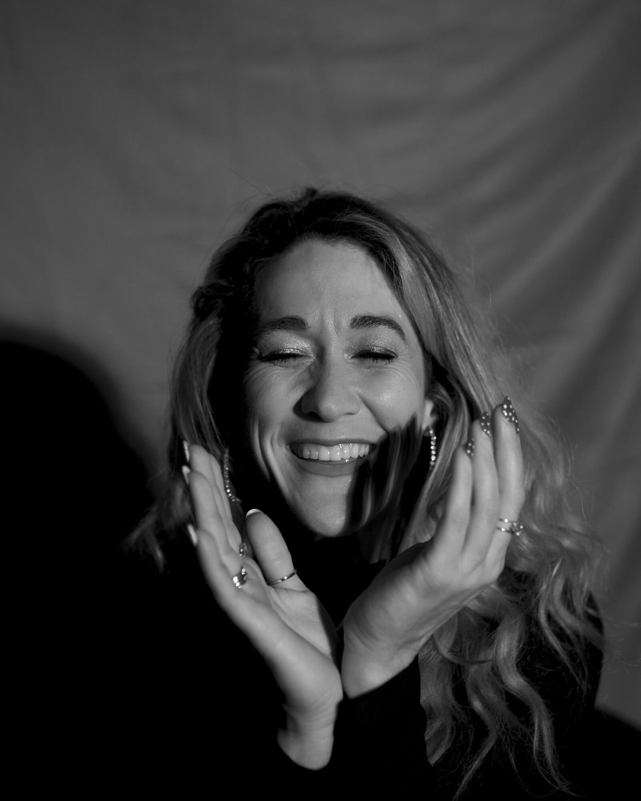 Tight black and white portrait, eyes closed mid-laugh, hands framing the face