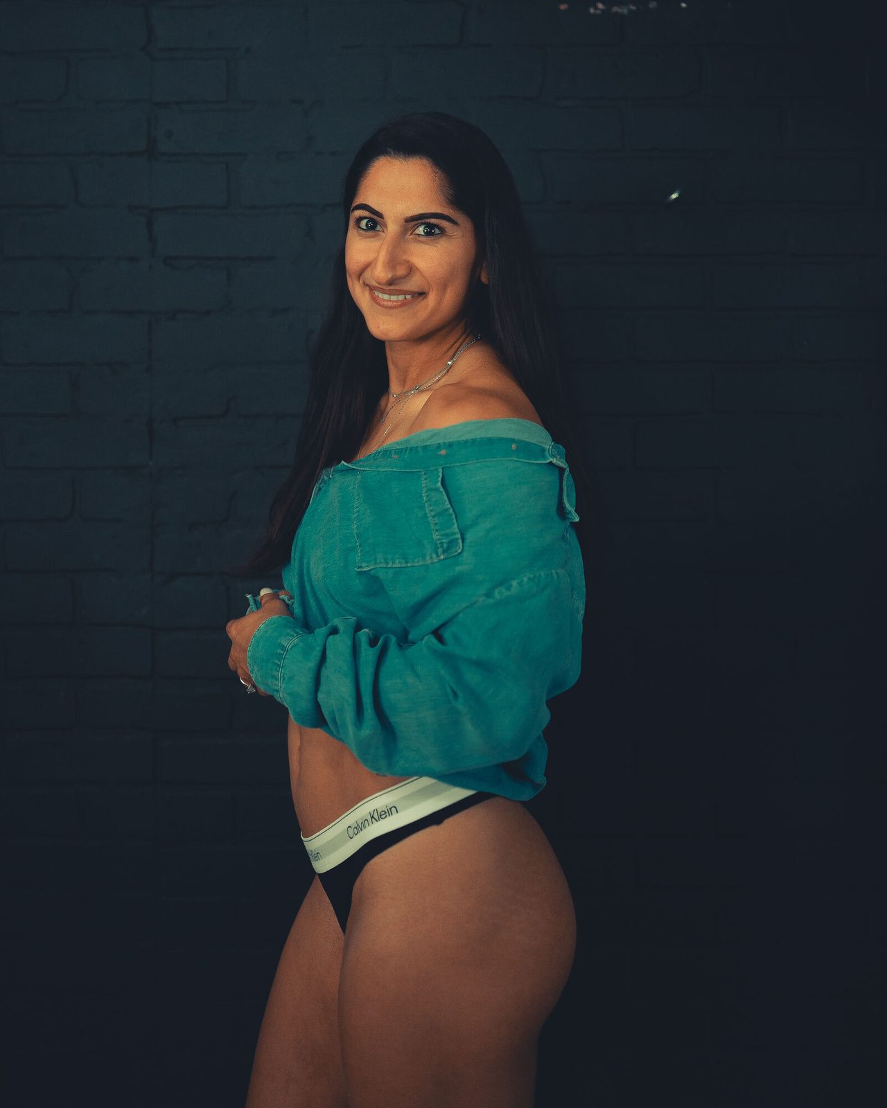Teal off-shoulder shirt in side profile against black brick